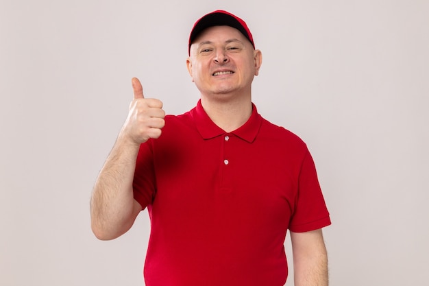 Happy delivery man in red uniform and cap looking at camera smiling cheerfully showing thumbs up standing over white background