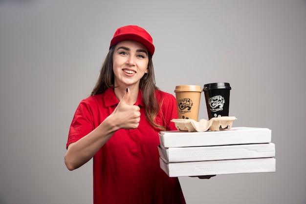 Happy delivery woman making thumbs up with pizza and coffee cups on gray wall.