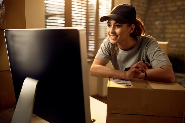 Счастливая женщина-доставщик, использующая компьютер в офисе во время работы с пакетами в офисе