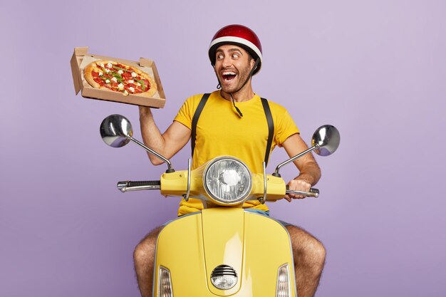 Happy deliveryman driving yellow scooter while holding pizza box