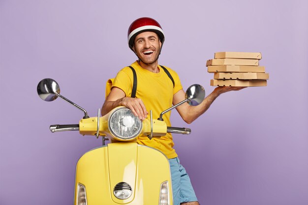 Happy deliveryman driving yellow scooter while holding pizza boxes