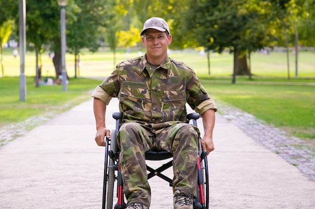 Счастливый военный-инвалид в инвалидной коляске в камуфляжной форме, двигаясь по тропинке в городском парке. Передний план. Ветеран войны или концепция инвалидности