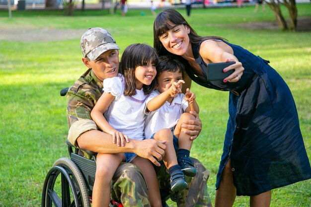 Счастливый инвалид отставной военный, держа детей на руках, пока его жена делает семейное селфи на мобильном телефоне. Ветеран войны или концепция возвращения домой
