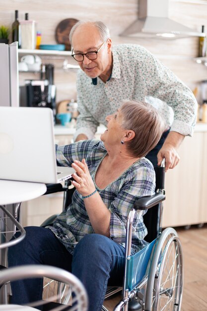 車椅子の幸せな障害者の年配の女性と夫が台所で話したり笑ったりしているラップトップでビデオ通話中に挨拶します。現代の通信オンラインインターネットウェブ技術を使用して麻痺した人