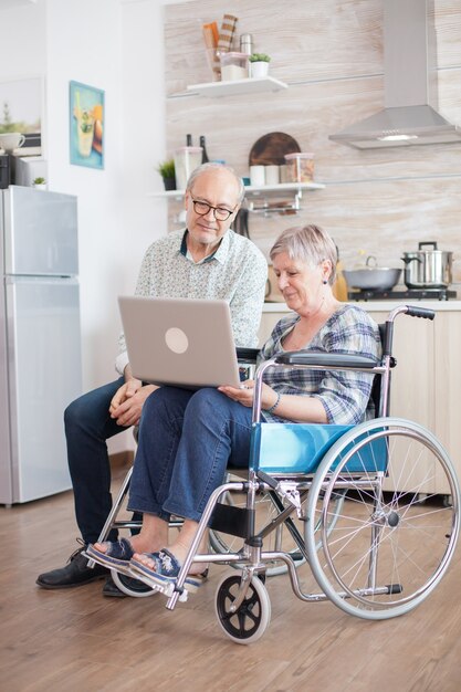 부엌에서 화상 회의를 하는 동안 행복한 장애인 여성. 휠체어를 탄 장애인 노인과 남편이 부엌에서 태블릿 PC로 화상 회의를 하고 있습니다. 마비된 노부와 그녀의 남편