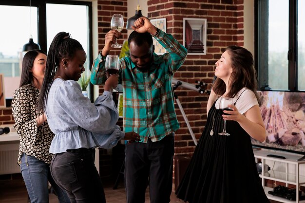 Happy diverse people listening to funky disco music while drinking wine and having fun time together. Multiethnic group of close friends dancing at party while celebrating important event.