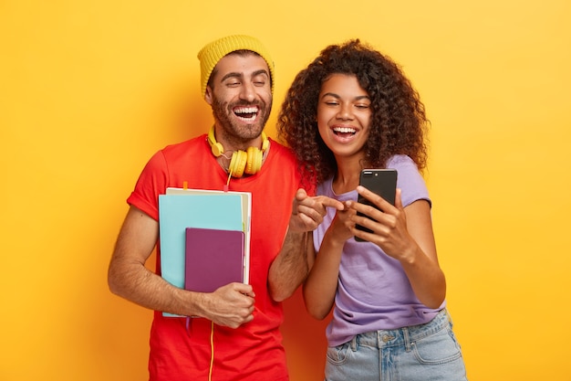 Happy diverse students look happily at smartphone device, hold notepad, wear stylish bright clothes