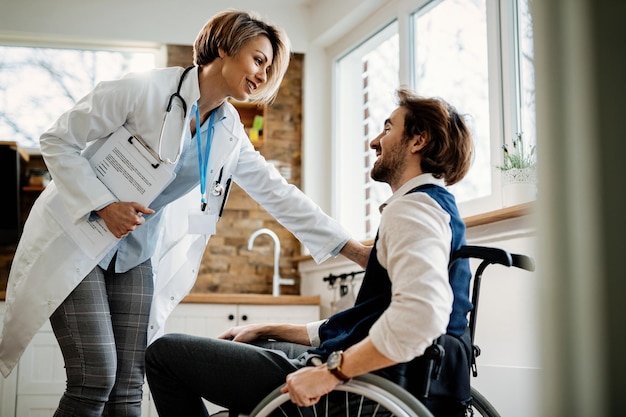 Happy doctor encouraging young man in wheelchair while talking to him during home visit.