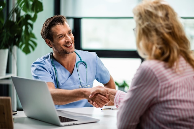 Happy doctor shaking hands with wife of his senior patient at medical clinic