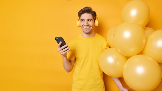 Happy dreamy man looks away with cheerful expression listens smartphone andstereo headphones for listening music holds bunch of balloons isolated over yellow background copy space for your promotion