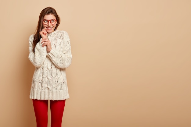 Happy dreamy woman has straight hair, looks glad fully with thoughtful expression, imagines positive moment in life, wears white jumper, red leggings, isolated over beige wall with free space