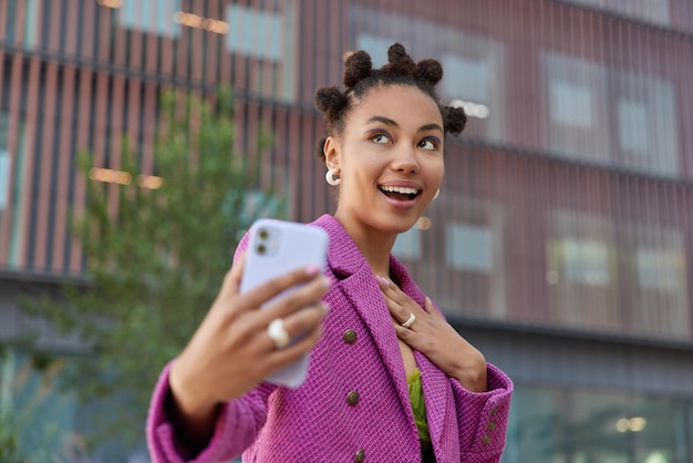 Happy dreamy woman with funny hairstyle makes photo of herself wears fashionable pink jacket looks somewhere positively poses outdoors against blurred background. Technology and urban lifestyle