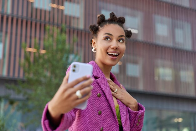 面白い髪型の幸せな夢のような女性は、自分の写真をファッショナブルなピンクのジャケットを着て、ぼやけた背景に対して屋外で積極的にポーズをとっています。テクノロジーと都会のライフスタイル
