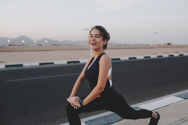 Happy early morning moments of hardworking motivated amazing young woman in sportswear stretching on road in tropical country. Training, workout, healthy lifestyle, smiling.