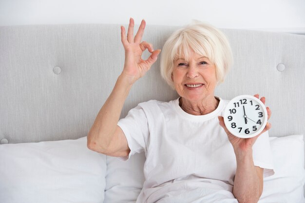 Happy elder woman holding a clock in her bed