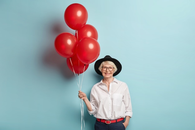 Happy elderly female in festive clothes, holds bunch of air balloons, celebrates anniversary, waits for children and guests, enjoys celebration, poses over blue wall. Pensioner on party