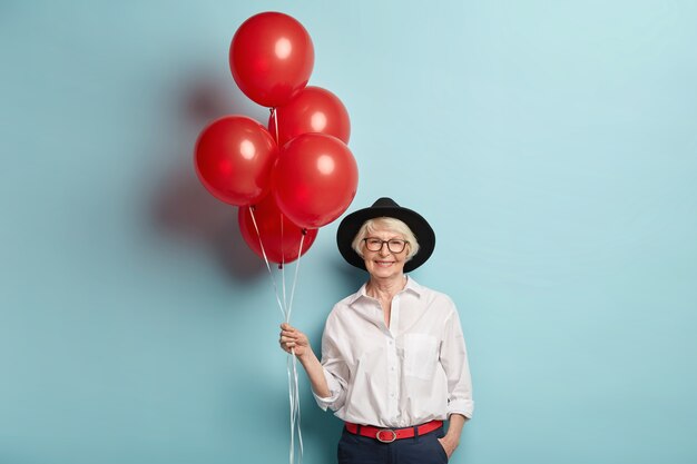 Бесплатное фото Счастливая пожилая женщина в праздничной одежде, держит кучу воздушных шаров, празднует годовщину, ждет детей и гостей, наслаждается празднованием, позирует над синей стеной. пенсионер на вечеринке