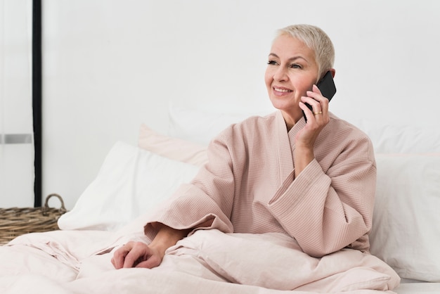 Happy elderly woman in bathrobe talking on smartphone in bed