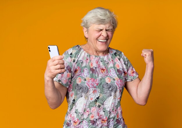 Happy elderly woman raises fist holding phone isolated on orange wall