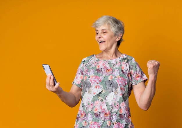 Happy elderly woman raises fist and looks at phone isolated on orange wall