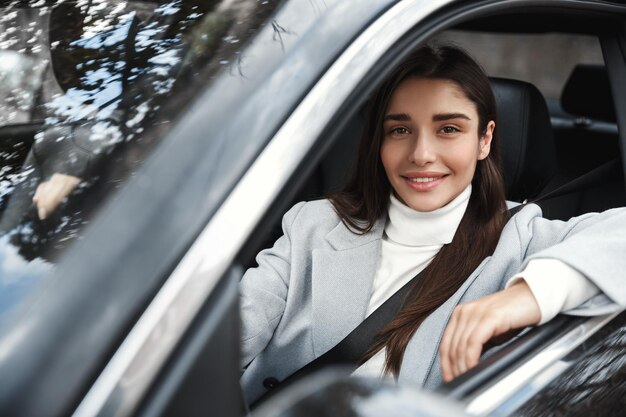 仕事で運転している固定シートベルトで車に座っている幸せなエレガントな女性窓の外を見てカメラに微笑んでいる女性ドライバー