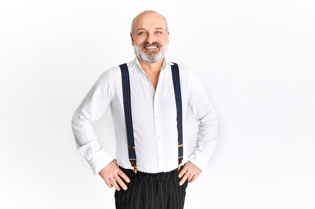 Happy emotional elderly man with gray beard and bald head rejoicing, holding hands on his waist, smiling cheerfully, posing against white background with copy space for your text