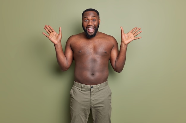 Free photo happy emotional man with dark skin raises palms reacts happily on unexpected surprise smiles broadly stands shirtless