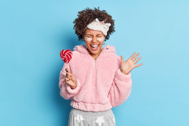 Happy emotional young Afro American girl exclaims from joy raises palm reacts on amazing news dressed in nightwear holds sweet candy on stick isolated over blue wall