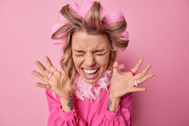 Happy emotional young woman keeps palms raised shouts loudly reacts on something amazing wears hair rollers for making curly hairstyle gets ready for date isolated over pink studio background