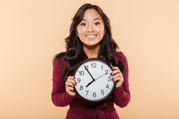 Happy emotions of asian woman with curly long hair holding clock showing nearly 8 waiting for something pleasant over peach background