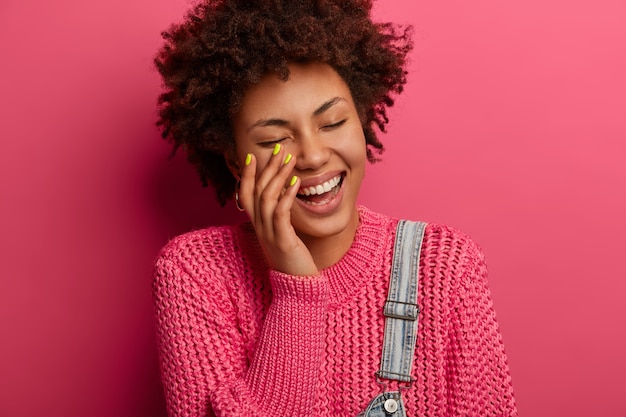 Foto gratuita concetto di emozioni e sentimenti felici. la donna afroamericana riccia gioiosa ride fuori dall'ascoltare scherzo esilarante, sorride ampiamente, essendo intrattenuta da un amico divertente, vestita casualmente, pose al coperto