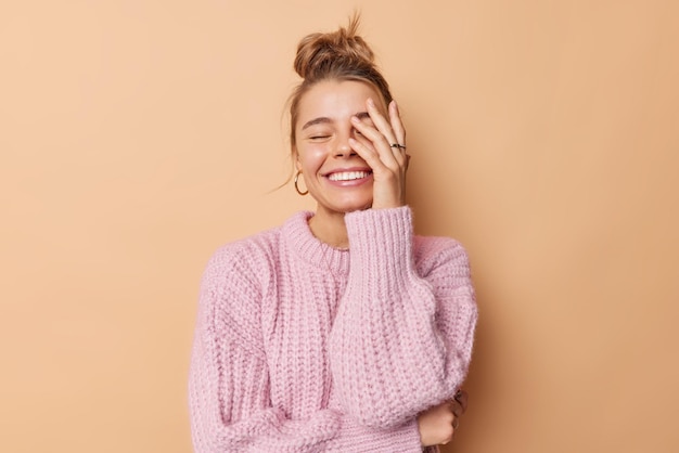 幸せな感情と感情の概念。髪をとかしたうれしそうな若い女性は、顔の手のひらを笑顔にします。ベージュの背景の上に分離されたニットのセーターを歯を見せて着ています。
