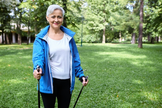 Pensionato femminile attivo energico felice in giacca blu che gode del nordic walking utilizzando pali appositamente progettati, respirando aria fresca all'aperto. attività fisica, stile di vita sano, persone e invecchiamento