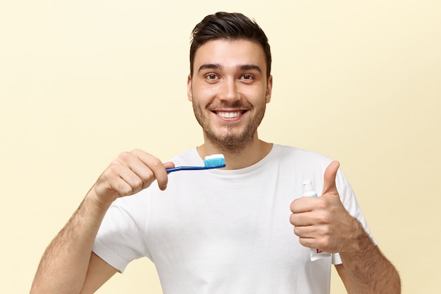 Happy energetic young European guy with stubble holding tooth brush with whitening paste and showing thumbs up gesture being in good mood.