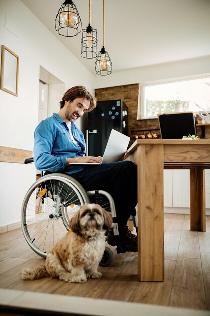휠체어를 탄 행복한 기업가는 집에서 개와 함께 노트북 작업을 합니다.