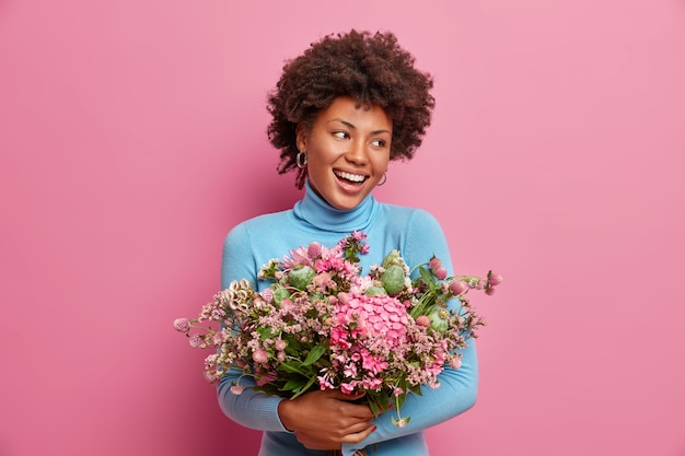 Happy ethnic Afro American woman embraces big bouquet of flowers, smiles broadly