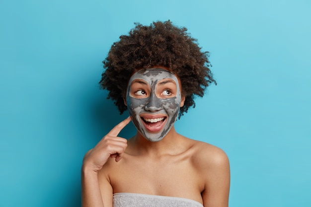 Happy ethnic curly haired woman smiles pleasantly applies facial clay mask wants to look beautiful wrapped in soft towel isolated over blue studio wall.