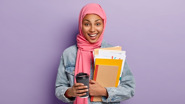 Happy ethnic female student has coffee break, holds takeaway cup of drink, carries notebook and papers, has pink scarf on head, Islamic religious views, poses indoor. People, culture, tradition