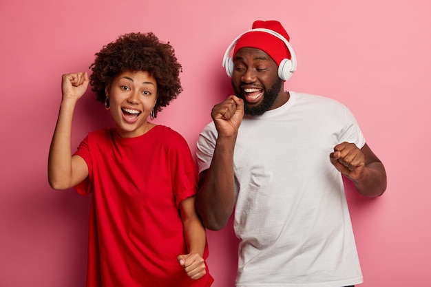 Счастливые этнические миллениал, девушка и парень двигаются в такт мелодии, слушают любимую музыку, имеют хорошее настроение, носят повседневные футболки. Современные технологии, свободное время и радость.