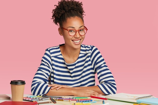 Happy ethnic student designer draws sketch for university exam, wears striped jacket and glasses