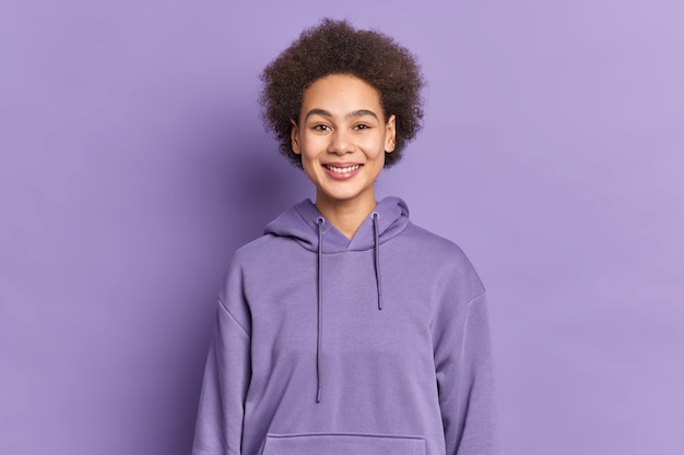 Happy ethnic teenager with Afro hair smiles positively wears purple hoodie being in good mood.