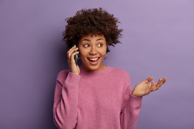 Happy ethnic woman talks on phone, looks away and gestures, discusses with impression her visit to theatre, dressed in casual jumper, concentrated aside, has toothy smile isolated on purple wall