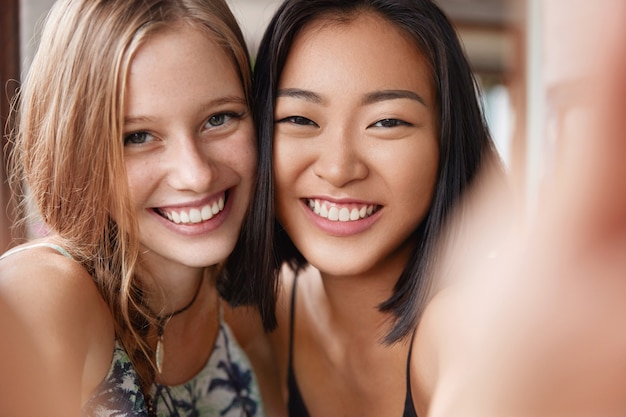 Happy European female embraces best Asian friend, make selfie, stand close to each other.