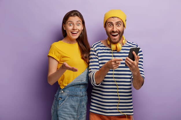 행복한 유럽 여성과 그녀의 남자 친구는 온라인 채팅에서 문자 메시지를 보내기 위해 현대 가제트를 사용하고 기쁜 표정으로 보며 스테레오 헤드폰을 사용합니다.