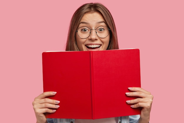 Happy European female student in spectacles, has positive expression, holds red book, rejoices successfully passed exam at university, isolated over pink wall. People, learning, reading