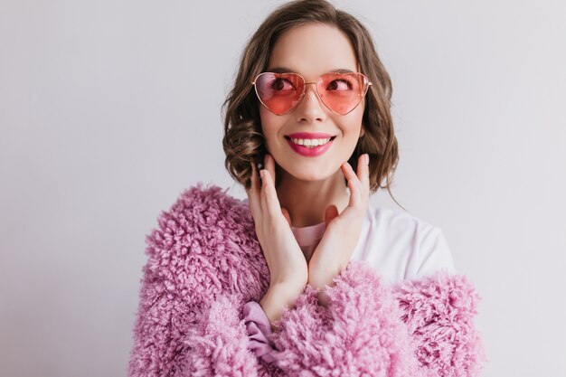  happy european girl in funny sunglasses isolated on white wall. Enchanting curly woman in pink fluffy jacket expressing positive emotions.