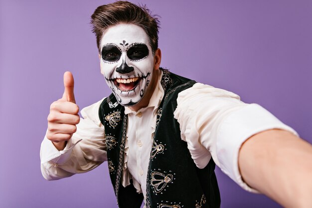 Счастливый европейский парень с традиционным мексиканским боди-артом позирует на фиолетовой стене. Стильный мужчина с макияжем зомби, делая селфи.