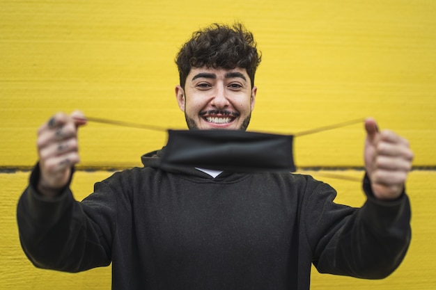 Happy European male showing a face mask on vibrant yellow wall