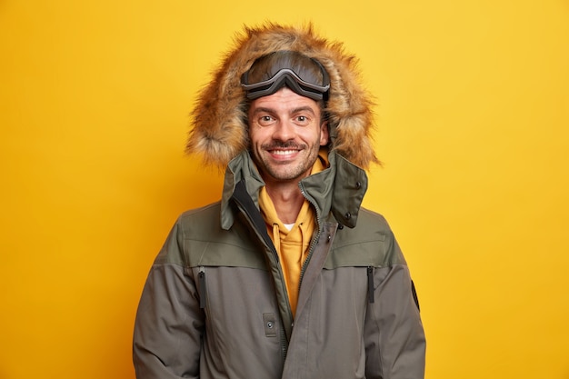 Free photo happy european man in jacket with fur hood feels warm and comfortable during winter time enjoys favorite season smiles happily wears skateboarding goggles has active rest.