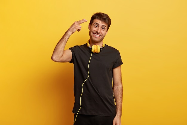 Happy European man shoots in temple, wears casual black t shirt, wears headphones on neck, tilts head